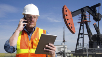 A royalty free image from the oil and gas industry of an oil engineer at a pumpjack using a tablet PC and cellular.