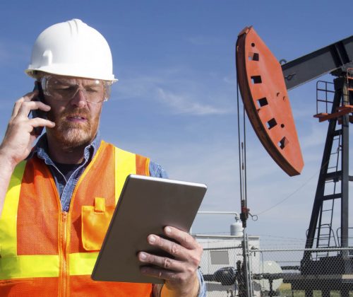 A royalty free image from the oil and gas industry of an oil engineer at a pumpjack using a tablet PC and cellular.